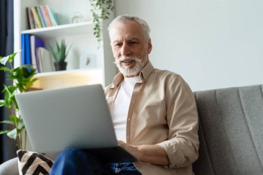 Mutlu emekli adam evdeki bir kanepede oturuyor, online iletişim ve modern teknoloji için bir dizüstü bilgisayarın rahatlığının tadını çıkarıyor.