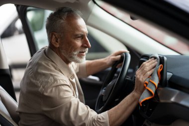 Yaşlı bir adam, aracının ön panelini titizlikle temizliyor. İçi tertemiz ve bakımlı olsun diye mikrofiber bir bez kullanıyor.
