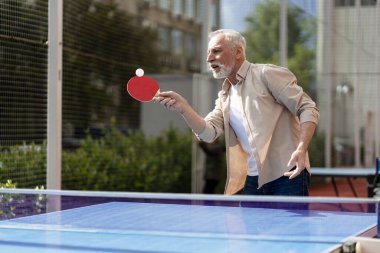 Odaklanmış son sınıf öğrencisi açık havada masa tenisi oynuyor güneşli bir günde topa kürekle vuruyor.