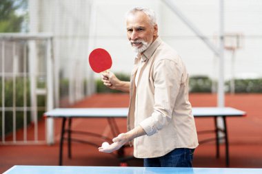 Konsantre olmuş yaşlı adam raket ve top tutuyor dışarıda masa tenisi oynarken servis yapmaya hazırlanıyor.