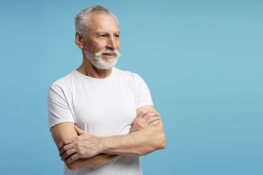 Mavi bir arka planda kollarını kavuştururken gülümseyen ve başka tarafa bakan kendinden emin bir adamın stüdyo portresi.
