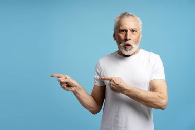 Beyaz saçlı, sakallı, beyaz tişörtlü yaşlı bir adam mavi arka planda her iki işaret parmağıyla işaret ediyor.