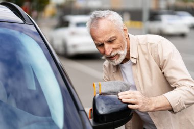 Yaşlı bir adam kalabalık bir caddede arabasının yan aynasını titizlikle cilalıyor, araba bakımı ve bakımının önemini vurguluyor.