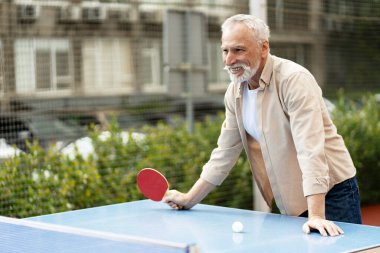Mutlu emekli adam güneşli bir günde pinpon maçından zevk alıyor, aktif yaşlanma ve sağlıklı bir yaşam tarzını teşvik ediyor.