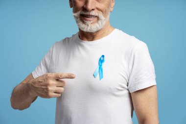 Mavi kurdelesini gösteren gülümseyen son sınıf öğrencisi beyaz tişörtüne iğnelenmiş, mavi arka planda prostat kanseri farkındalığını destekliyor.