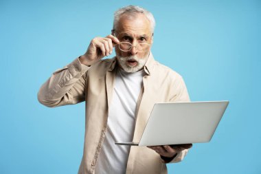Beyaz sakallı, saçları olan, gözlüklü ve elinde mavi arka planda şok olmuş yüz ifadesi olan dizüstü bilgisayarı olan olgun bir adam.