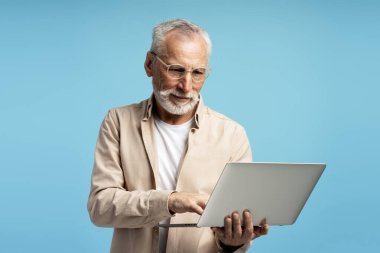 Odaklanmış olgun öğretmen dizüstü bilgisayar kullanıyor, internette çalışıyor, öğreniyor, mavi stüdyo arka planında tek başına duruyor.