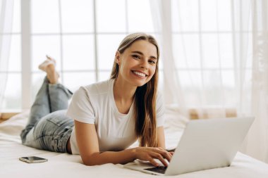 Yatakta rahat bir şekilde uzanan genç bir kadın dizüstü bilgisayar kullanarak çalışıyor ve gülümsüyor. Çevrimiçi teknoloji kavramı