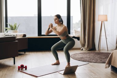 Kulaklık takan sağlıklı kadın evde dizüstü bilgisayar kullanarak online fitness dersini takip ediyor. Eğitim kavramı