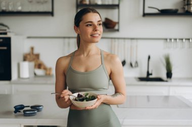 Sportif kadın, güzel sporcu kadın modern mutfakta sağlıklı salata yiyor ve başka tarafa bakıyor. Yiyecek konsepti