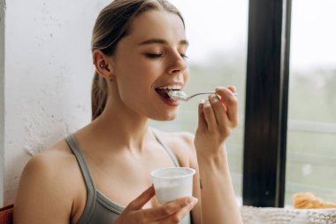 Antrenmandan sonra elinde kaşıkla yoğurt yiyen gülümseyen sporcu kadın. Sağlık yaşam tarzı kavramı
