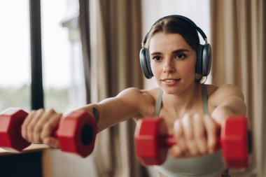 Oturma odasında halterlerle egzersiz yapan ve kulaklıklarla müzik dinleyen sağlıklı bir kadın. Spor, antrenman konsepti