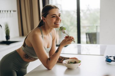 Sporcu kadın, spor kıyafetleri giyen genç kadın antrenmandan sonra mutfakta salata yiyor. Sağlık hizmeti kavramı