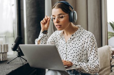 Genç bir kadın kulaklık, dizüstü bilgisayar ve mikrofon kullanarak stüdyodan podcast yapıyor. Çevrimiçi iletişim kavramı