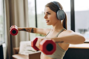 Fit bir kadın evde müzik dinlerken dambıllarla dikey yükseliş yapıyor. Eğitim kavramı