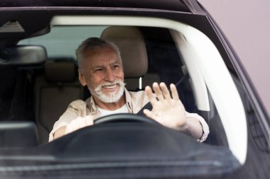 Gülümseyen kıdemli bir adam araba kullanıyor, pencereden arkadaşına ya da ailesine el sallıyor, yolculuğunda neşe ve sıcaklık saçıyor.