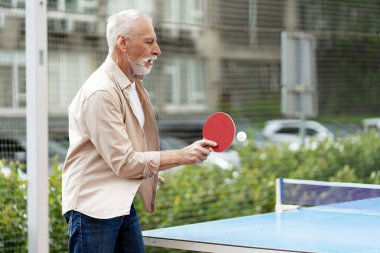 Beyaz saçlı ve sakallı yaşlı adam dışarıda pinpon oynuyor topa odaklanarak vuruyor ve hareketli bir şehir parkında aktif emeklilik hayatının tadını çıkarıyor.