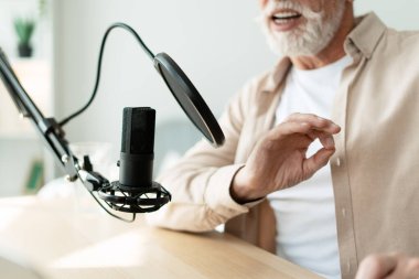 Yaşlı bir adam ses içeriğini kaydediyor, pop filtresiyle mikrofona konuşuyor, rahat stüdyosundan canlı yayın bölümleri ya da internet radyo programları yaratıyor.