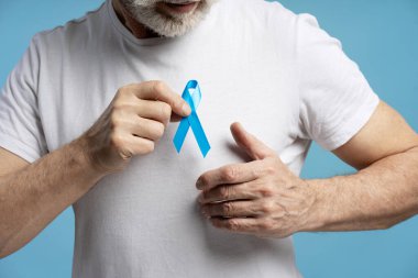 Göğsünde açık mavi kurdele tutan kıdemli bir adamın yakın çekimi prostat kanseri farkındalığını, erken teşhisi ve erkek sağlığını sembolize ediyor.