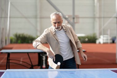 Konsantre olmuş yaşlı adam dışarıda masa tenisi oynuyor güneşli bir günde topa kürekle vuruyor aktif yaşam tarzını sürdürüyor ve emekliliğin tadını çıkarıyor. Rekabet konsepti
