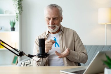 Prostat kanseri farkındalığının sembolü olan mavi kurdele takan gri saçlı olgun bir adam profesyonel mikrofon ve dizüstü bilgisayar kullanarak erkeklerin sağlığı hakkında podcast yapıyor. Tedavi kavramı