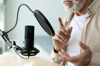 Yaşlı radyo sunucusu mikrofonla konuşuyor, podcast ya da röportaj yapıyor, el kol hareketi yapıyor, profesyonel mikrofon kullanıyor, kayıt stüdyosunda ya da ev stüdyosunda. Çevrimiçi teknoloji kavramı
