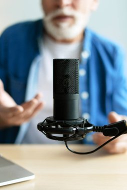 Sakallı kıdemli bir adam elle işaret ederken ses içeriğini kaydediyor, bir dizüstü bilgisayar ve profesyonel yoğunlaştırıcı mikrofon ile donatılmış bir masada oturuyor. Çevrimiçi iletişim kavramı