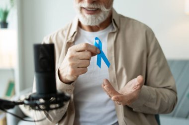 Evde mikrofon kullanarak podcast yaparken prostat kanseri farkındalığının sembolü olarak elinde mavi kurdele tutan gri sakallı son sınıf öğrencisi gülümsüyor. Sağlık hizmeti kavramı