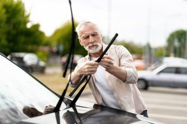 Sakallı kıdemli bir adam otoparkta park etmiş arabasının ön cam sileceklerini titizlikle değiştiriyor. Aracın işlevselliğini ve güvenliğini sağlarken bakımını yapıyor. Onarım kavramı