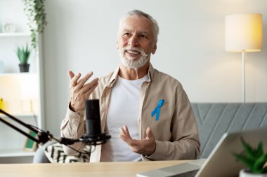 Gri saçlı, sakallı, mavi bilinç kurdelesi takmış, podcast bölümünü kaydederken mikrofona konuşan, elleriyle hareket eden ve gülümseyen olgun bir adam. Sağlık hizmeti kavramı