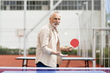 Gri saçlı ve sakallı, sıradan kıyafetler giyen yaşlı bir adam açık masa tenisi masasında pinpon topu servis ediyor, aktif yaşam tarzını ve spora olan tutkusunu gösteriyor. Etkin yaşam tarzı kavramı