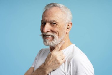 Beyaz sakallı ve gri saçlı mutlu emekli adam mavi arka planda gülümserken işitme cihazını işaret ediyor, yaşlılar için işitme ve asistan teknolojisini destekliyor. Tedavi kavramı