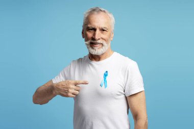 Beyaz tişörtüne takılmış mavi kurdeleyi işaret eden kıdemli bir adam prostat kanseri ve erkek sağlığı bilincini artırıyor, açık mavi arka planda erken teşhisi ve desteği teşvik ediyor.