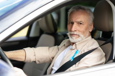 Sakallı son sınıf öğrencisi, prostat kanseri farkındalığı ayı için açık mavi kurdele takıyor, emniyet kemerine bağlanmış, izleyiciye bakıyor. Sağlık hizmeti kavramı