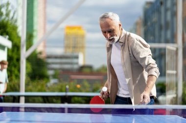Güneşli bir günde dışarıda masa tenisi oynayan, şehir hayatı ve enerjisiyle dolu bir ortamda aktif kalarak emekliliğinin tadını çıkaran konsantre yaşlı bir adam. Spor konsepti