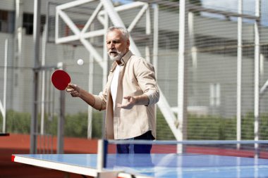 Konsantre olmuş yaşlı bir adam açık havada masa tenisi oynuyor güneşli bir günde kürekle topa vuruyor aktif emekliliğin ve sağlıklı yaşam tarzının tadını çıkarıyor. Tenis konsepti