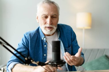 Gri saçlı ve sakallı yaşlı bir adam ev stüdyosunda ses yayını yapıyor, profesyonel bir mikrofon kullanıyor ve sezgilerini paylaşırken kendini ifade ediyor.