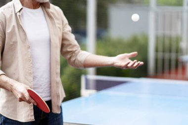 Ping pong raketi tutan yaşlı bir adam, açık masa tenisi masasında topu servis etmeye hazırlanıyor, canlı bir bahçe ortamında dostça bir maç başlatıyor. Spor konsepti