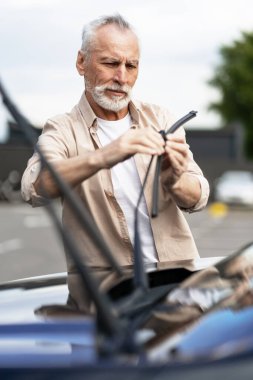 Odaklanmış kıdemli bir adam arabasına yeni silecekler yerleştiriyor ve hava koşullarında güvenli sürüş için net bir görüş sağlıyor. Servis kavramı