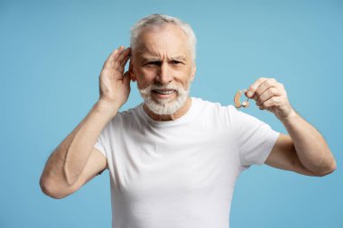 Beyaz sakallı, beyaz tişörtlü, işitme cihazı tutan ve mavi arka planda duyma güçlüğü çeken yaşlı bir adam. Sağlık hizmeti kavramı