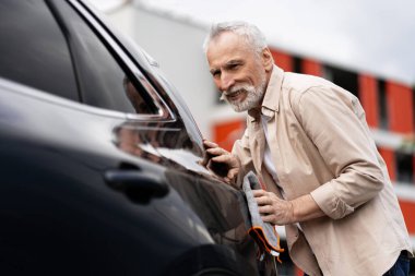 Gülümseyen kıdemli bir adam yeni siyah arabasını mikrofiber bezle cilalıyor, araba boyasıyla ilgileniyor, satın almasından memnun, araba detaylandırması konseptiyle uğraşıyor.