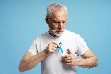 Destekleyici kıdemli adam beyaz tişörte mavi kurdele takıyor, dayanışma gösteriyor ve prostat kanseri ve erkek sağlığı sorunları için farkındalığı artırıyor