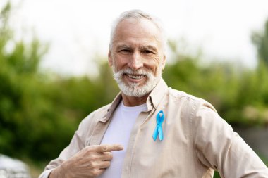 Mutlu, gülümseyen son sınıf öğrencisi gururla gömleğindeki mavi kurdeleyi işaret ederek, açık havada güneşli bir günün tadını çıkarırken prostat kanseri bilinçlendirme kampanyasını destekliyor.