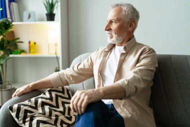 Mutlu bir emekli adam rahat bir oturma odasında rahat bir şekilde oturup, pencereden dışarı dikkatlice bakarken huzurlu bir rahatlama anının tadını çıkarıyor, emeklilikten memnuniyeti kucaklıyor.