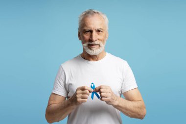 Elinde mavi kurdele tutan gülümseyen bir kıdemli adamın portresi, prostat kanseri, kolon kanseri ve kronik yorgunluk sendromu için farkındalığı sembolize ediyor.