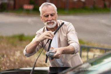 Sakallı kıdemli bir adam titizlikle araba sileceklerini mikrofiber bezle temizliyor, görüş mesafesini arttırıyor ve yolda güvenli bir sürüş deneyimi sağlıyor. Araba yıkama konsepti