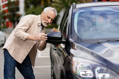 Yaşlı adam dikkatli bir şekilde arabasının yan aynasını ayarlıyor, parlak ve güneşli bir günde güvenli sürüş için ideal konumu sağlıyor, sorumlu araç bakımı ve güvenlik farkındalığını gösteriyor