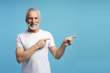 Beyaz sakallı, beyaz tişörtlü, mutlu emekli adam mavi stüdyo arka planında boş fotokopi alanı olduğunu işaret ediyor. Reklam kavramı
