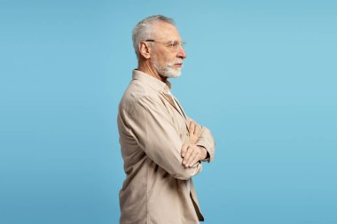 Gözlük ve bej gömlek takmış, bilgelik ve deneyimin bir görüntüsünü yansıtan, mavi arka plan karşısında duran kendinden emin kıdemli bir adamın profili.