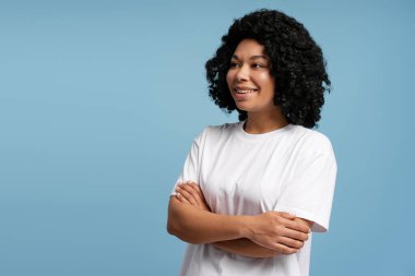 Doğal kıvırcık saçlı, gülümseyen genç bir kadın mavi arka planda kollarını kavuşturarak poz veriyor. Yaşam tarzı, çeşitlilik ve duygu kavramı